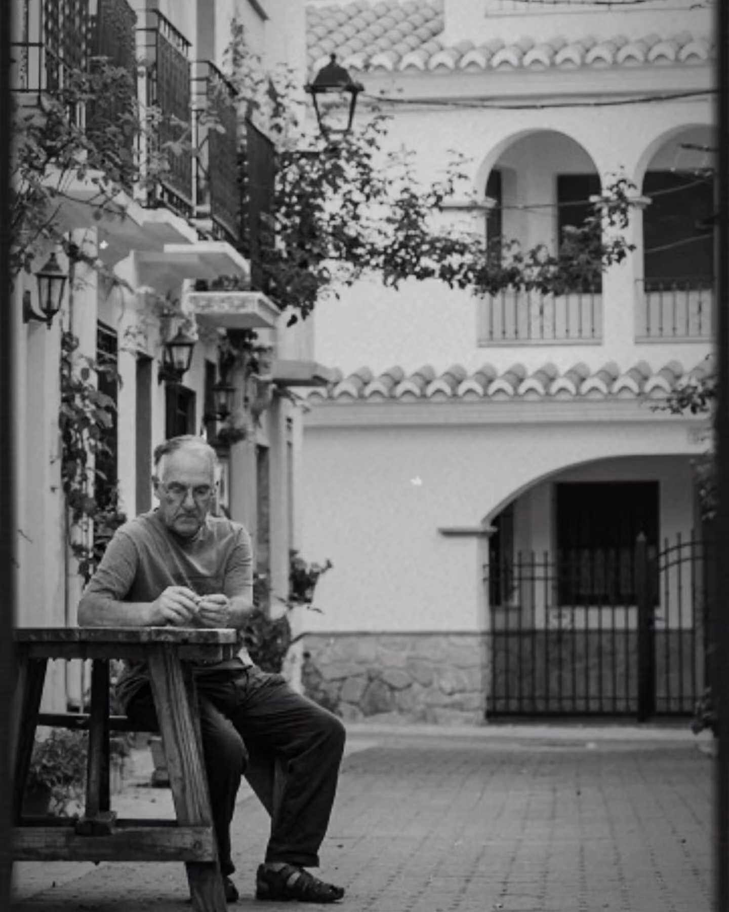 Fotografía en blanco y negro de un hombre mayor sentado a una mesa bajo los soportales de una plaza del Alto Turia.